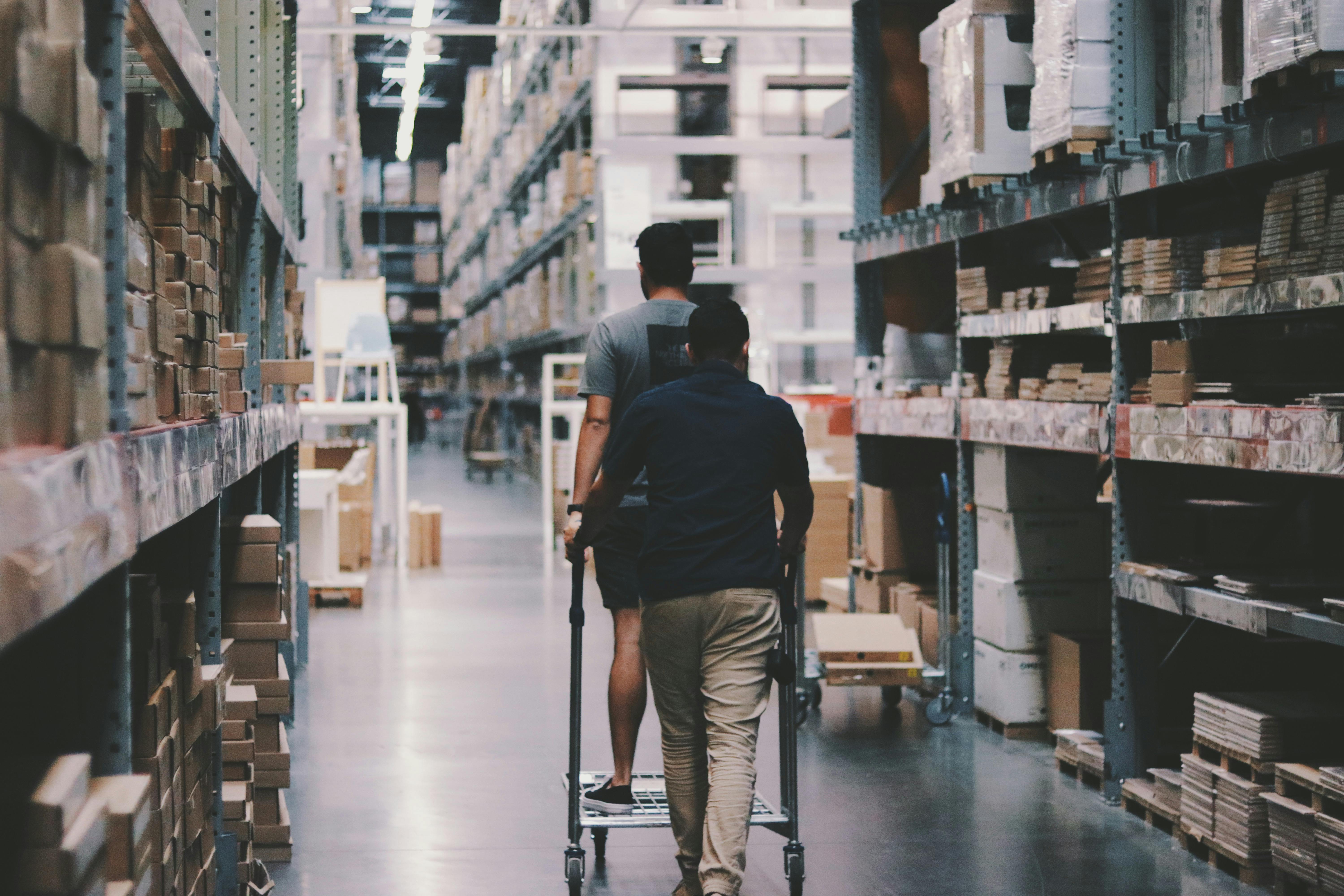 Employés poussant un chariot dans une allée d’un entrepôt logistique rempli de cartons et palettes