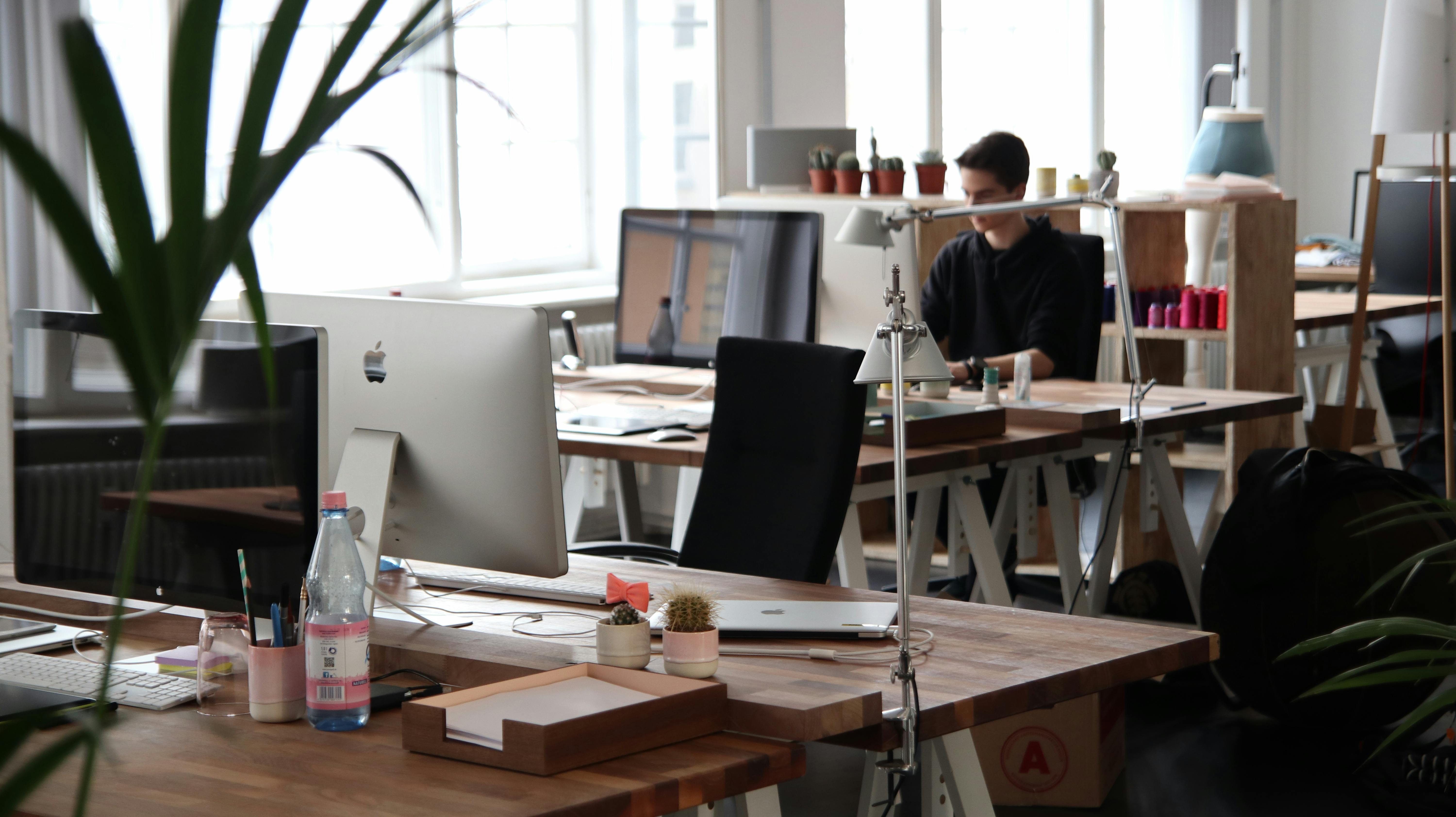 Espace de travail collaboratif moderne avec bureaux en bois, écrans Apple, plantes vertes et lumière naturelle, dans un open space design et lumineux.