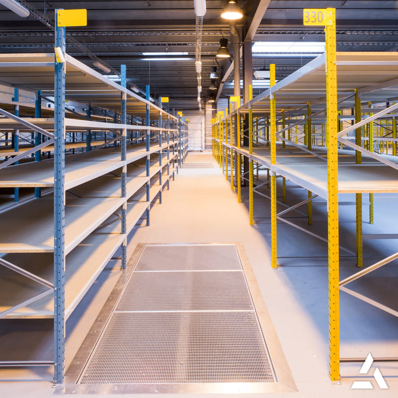 Rayonnages métalliques industriels vides en entrepôt logistique, allée centrale avec éclairage LED et montants jaunes.