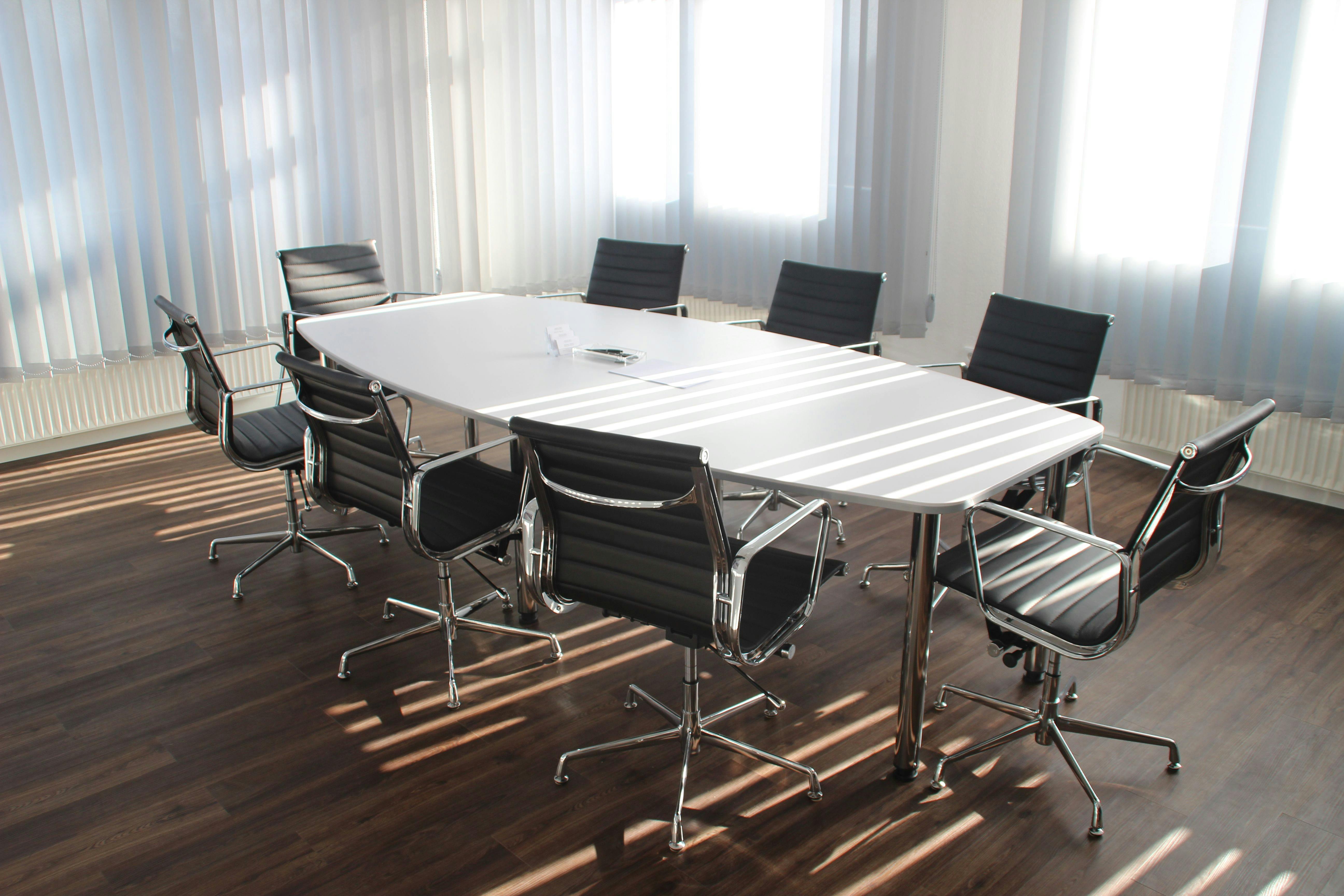 Salle de réunion moderne avec table blanche et chaises de bureau ergonomiques noires sur sol en bois foncé, éclairée par la lumière naturelle à travers des stores vénitiens.